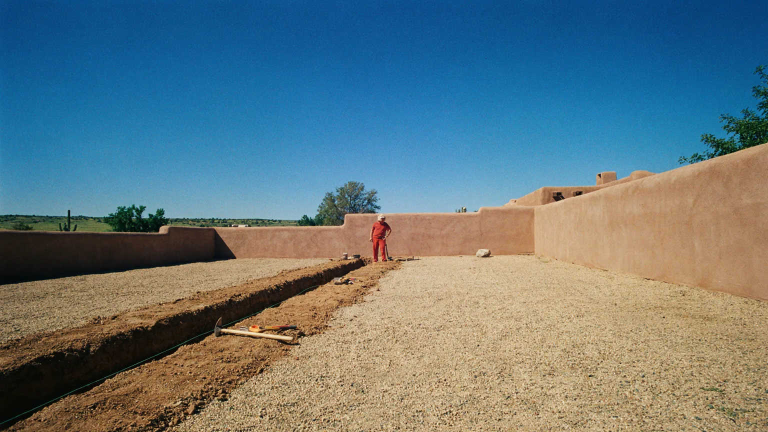An irrigation trench crossing a gravel yard toward an adobe garden wall
